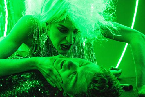 Ein komplett in grünes Licht getauchtes Foto der Schauspielerin Alberta von Poelnitz, wie sie sich über den am Boden liegenden Schauspieler Paul Behren beugt und ihn am Kinn packt