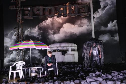 Der Schauspieler Paul Behren sitzt auf einem weißen Plastikstuhl unter einem bunten Sonnenschirm. Um ihn überall schwarze Kohle um ihn herum. Im Hintergrund dunkle Wolken am Himmel und ein gigantisches Schild, das verkehrt herum steht und von der anderen Seite den Schriftzug "Europe" zeigt.