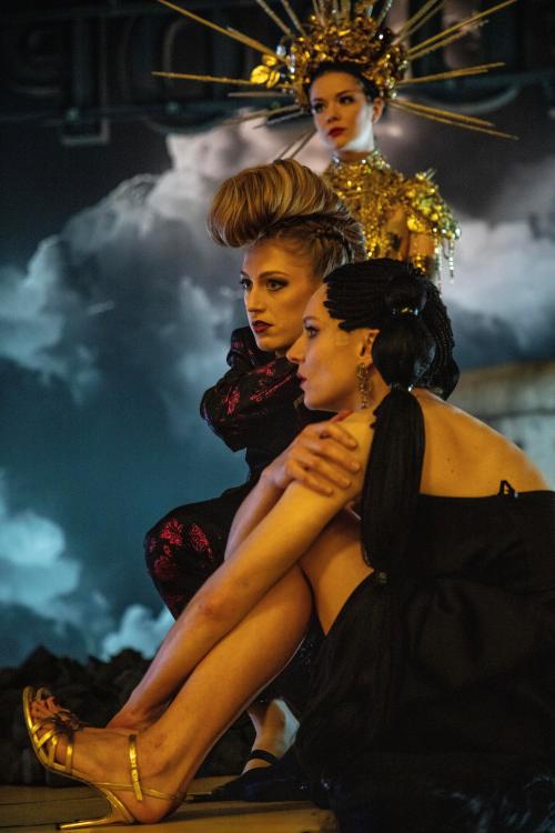 Vor einem dunklen, wolkenüberzogenen Hintergrund sitzen die Schauspielerinnen Lilith Stangenberg, Alberta von Poelnitz und Linn Reusse. Sie tragen dramatisches Make-Up, edle und große Kleider und aufwendigen Haarschmuck