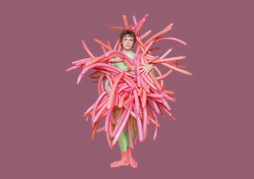 Ein Mann steht vor einem neutralen Hintergrund. Er ist mit bunten, aufgeblasenen Röhrenluftballons eingehült, die in alle Richtungen abstehen. Mit seinen Händen hält er die Luftballons in Position.