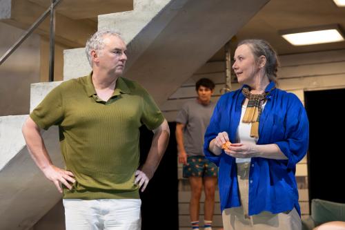 Hermann Book und Christine Ochsenhofer stehen nebeneinander vor einer steinernen Treppe und sehen sich an. Hermann hat die Hände in die Hüften gestemmt. Im Hintergrund steht Silvio Kretschmer, der die beiden beobachtet.