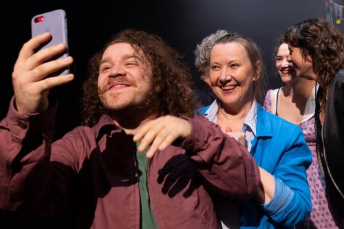 Nahaufnahme vom Ensemble. Parsa Yaghoubi Pour und Christine Ochsenhofer lächeln in ein Smartphone. Die anderen lächeln sich im Hintergrund an und stehen dabei nah beieinander.