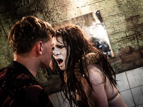 Paul Behren ist von der Seite zu sehen. Dicht vor seinem Gesicht ist Lilith Stangenberg, ihr Gesicht ist komplett weiß geschminkt. Sie schaut ihm in die Augen, ihr Mund ist weit geöffnet. Im Hintergrund ein Spiegel und eine grünlich-weiße Ziegelwand.