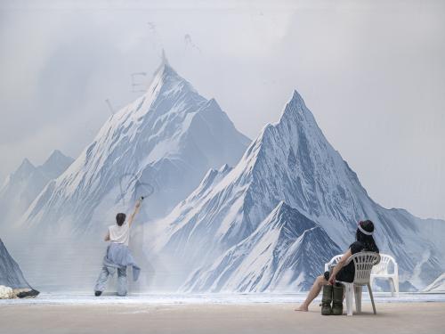 Sachiko Hara malt mit einem Pinsel ein Herz auf die schneebedeckte Berglandschaft. Sasha Rau sitzt etwas weiter von ihr entfernt auf einem Stuhl und schaut ihr dabei zu.