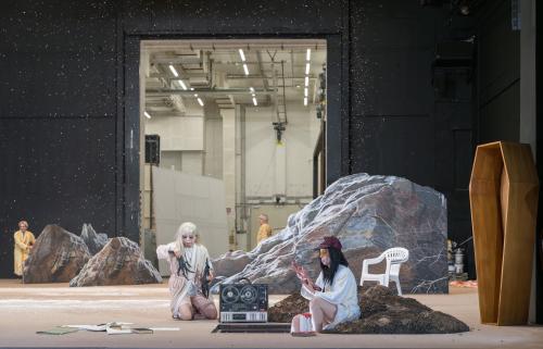 Sachiko Hara und Sasha Rau sitzen auf den Boden bei einem Loch im Boden. Sachiko Hara hält große Krähenfüße und Sacha Rau einen blutigen Arm in der Hand. Rechts ein senkrecht aufgestellter Sarg. Im Hintergrund sind große Felsen zu sehen. Weiße Schnipsel fallen von der Decke. Im Hintergrund eine schwarze Wand mit einem offenen Portal zur Hinterbühne.