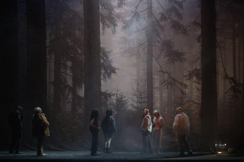 Das Ensemble steht verteilt auf der Bühne und schaut auf das Bühnenbild, das einen düsteren Wald abbildet.
