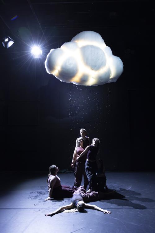 Sechs Tänzer*innen stehen, sitzen und liegen unter einer Wolke, die regnet. Einige schauen nach oben in die Wassertropfen.