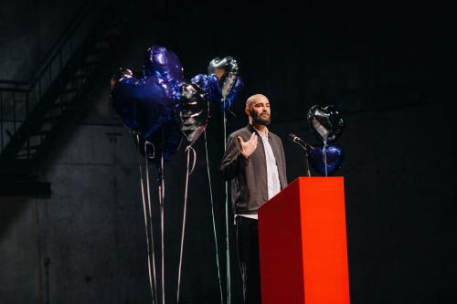 Philipp Staab steht an einem roten Podium, auf dem ein Mikrofon platziert ist, er gestikuliert mit der rechten Hand. Um ihn herum ist eine Ansammlung von silbernen und blauen herzförmigen Heliumluftballons.