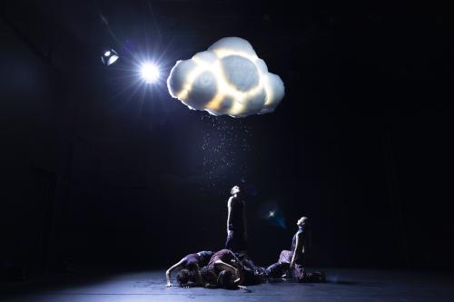 Einige Tänzer*innen stehen und krabbeln unter einer Wolke, die regnet. Einige schauen nach oben in die Wassertropfen.