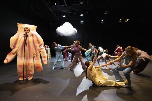 Es sind viele Tänzer*innen auf der Bühne in Bewegung. Viele tanzen in Zweierpaaren und berühren sich. Am Mikrofon steht ein Tänzer in einem Handkostüm. 