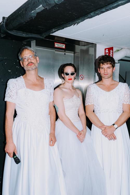 Martin Györffy, Christian Tschirner und lynn t musiol stehen in einer Reihe nebeneinander in langen weißen Kleidern. Sie posieren vor einem Fahrstuhl.