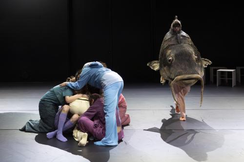 Eine Gruppe von Tänzer*innen bildet einen Menschenhaufen. Daneben läuft ein Tänzer in einem Fischkostüm vorbei. 