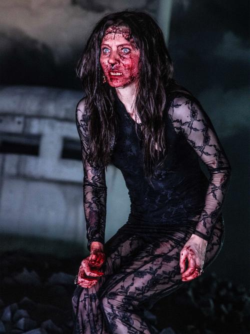 Lilith Stangenberg mit einer Perücke aus langen braunen Haaren. Ihr Gesicht ist vollständig mit Kunst blutbeschmiert, sie hat den Mund geöffnet und die Augen aufgerissen. Sie trägt ein schwarz-transparentes Oberteil.