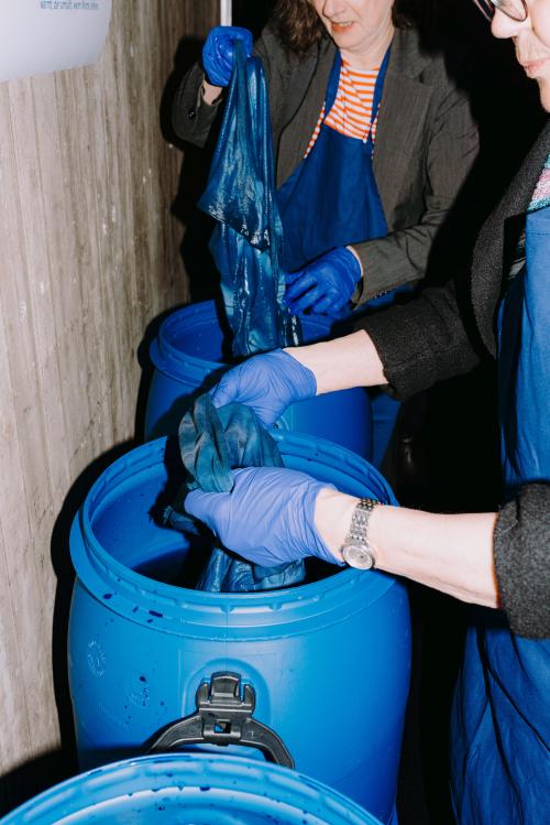 Zwei Personen tunken Tücher in drei große blaue Fässer mit blauer Farbe darin. Die Frauen tragen blaue Handschuhe und blaue Schürzen.