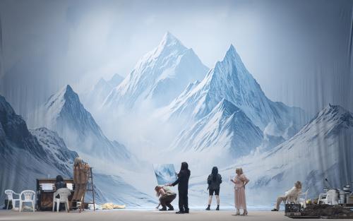 Das Ensemble steht verteilt auf der Bühne und schaut auf eine schneebedeckte Berglandschaft. Links steht ein Klavier und verschiedene Gegenstände sind auf dem Bühnenbild verteilt. Jemand hält ein Bild von der bergigen Landschaft in den Händen.