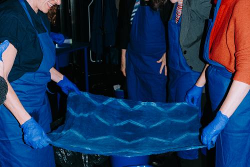 Zwei Personen in blauen Schürzen und mit blauen Handschuhen breiten gemeinsam ein blaues Tuch mit einem Muster aus. Im Hintergrund stehen weitere Personen in blauen Schürzen.