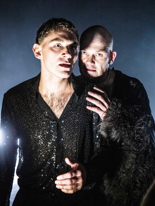 Paul Behren und Matti Krause in Nahaufnahme, ihre Gesichter sind hell beleuchtet mit starkem Schlagschatten. Sie tragen schwarze glitzernde Oberteile. Mattis Hand liegt auf Paul Behrens linker Schulter. Ihre Münder sind geöffnet, Matti Krause schaut in die Kamera.