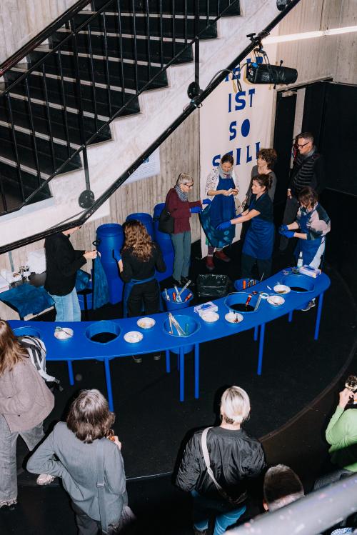 Viele Menschen stehen verteilt im MalerSaal Foyer, einige halten blaue Tücher in den Händen. In der Mitte des Raumes ist ein blauer langer Tisch, auf dem sich Werkzeuge und Bastelutensilien befinden. Es sind Eimer in den Tisch eingelassen.