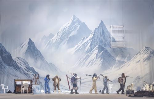 Das Ensemble läuft in einer Reihe über die Bühne. Alle tragen Skianzüge sowie Skier und Skistöcke in den Händen. Links steht ein Klavier und verschiedene Gegenstände sind auf dem Bühnenbild verteilt. Im Hintergrund eine schneebedeckte Berglandschaft.