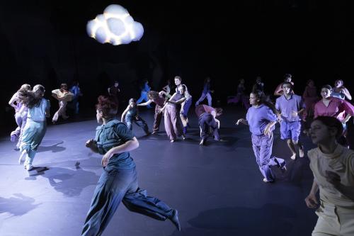 Die Tänzer*innen rennen auf der Bühne im Kreis. Über ihnen eine Wolke, die leicht leuchtet. 