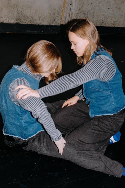 Lili Süper kniet auf dem Boden. Mit ihrem rechten Arm hält sie eine Puppe, die genau so aussieht wie sie (gleiche Größe, gleiche Kleidung, gleiche Haare).
