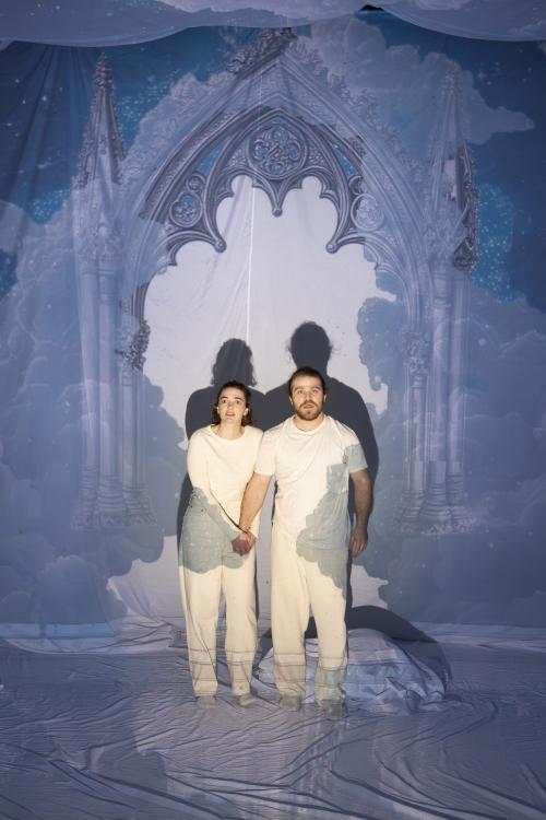 Victoria Kraft und Parsa Yaghoubi Pour stehen nebeneinander und blicken mit offenen Mündern in die Ferne. Victoria hält Parsas Hand mit beiden Händen umklammert. Sie stehen vor einem aufgespannten weißen Tuch, auf das ein gotischer Torbogen projiziert wird, der die beiden umringt. 