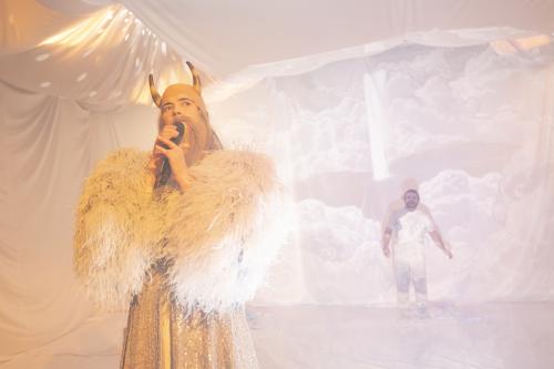 Victoria Kraft steht verkleidet mit einem langen weißen Bart, Felljacke und Hörnern auf dem Kopf im Vordergrund und hält ein Mikrofon in beiden Händen. Parsa Yaghoubi Pour ist im Hintergrund schwach zu erkennen. Beide befinden sich in einem Raum aus weißen Tüchern.