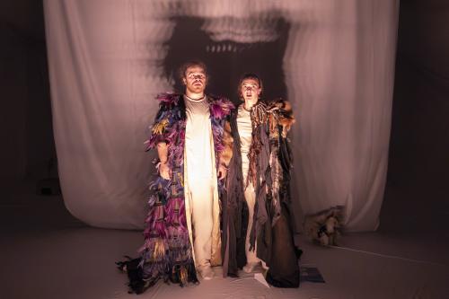 Parsa Yaghoubi Pour und Victoria Kraft stehen dicht nebeneinander und blicken mit offenen Mündern in die Ferne. Sie tragen beide lange Umhänge, Parsas mit bunten Federn und Victorias mit Tierfellen und kleinen Knochen bestückt.