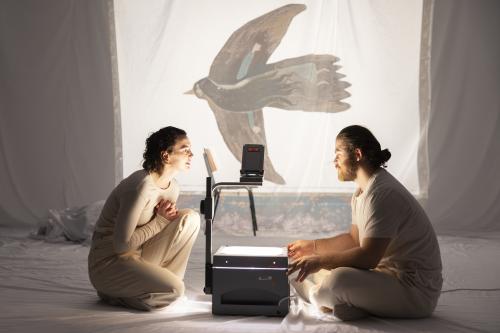 Victoria Kraft und Parsa Yaghoubi Pour sitzen sich gegenüber auf dem Boden. In ihrer Mitte steht ein Overheadprojektor, der auf ein weißes Tuch im Hintergrund ein gemaltes Bild von einem Vogel projiziert. Im Inneren des Vogels ist eine Frau mit langen Haaren gezeichnet.