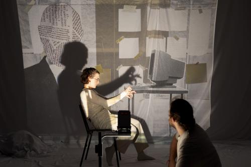 Victoria Kraft sitzt seitlich auf einem Stuhl und macht im Licht eines Overheadprojektors ein Schattenspiel mit ihren Händen. Parsa Yaghoubi Pour schaut ihr dabei zu. Das Bild, dass im Hintergrund auf die Leinwand projiziert wird, zeigt eine Bürowand, an der einige Zettel geklebt sind, und einen Tisch mit einem Computer. 