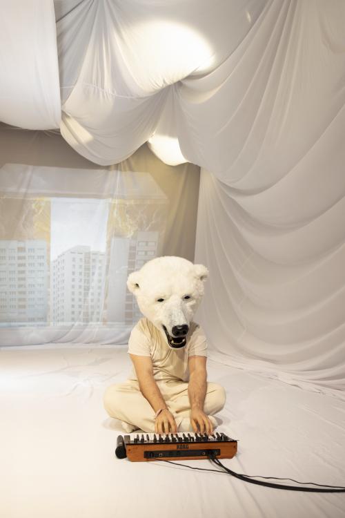 Parsa Yaghoubi Pour sitzt auf dem Boden im Schneidersitz und spielt auf einem kleinen Keyboard. Auf seinem Kopf trägt er eine große Eisbärenmaske. Um ihn herum sind weiße Tücher aufgespannt, die Falten schlagen.