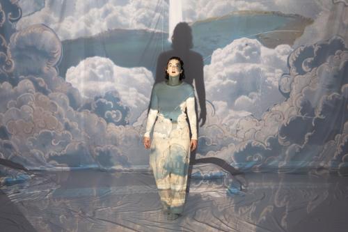 Victoria Kraft steht im Mittelpunkt und starrt mit weit aufgerissenen Augen und offenen Mund nach oben. Sie ist umgeben von weißen Tüchern, auf denen ein Wolkenbild projiziert wird.