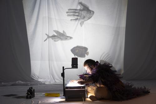 Parsa Yaghoubi Pour sitzt auf dem Boden und legt eine Folie auf einen Overheadprojektor. Das Bild, das dadurch an ein weißes Tuch im Hintergrund projiziert wird, zeigt zwei Fische und ein Gemisch aus Fisch und Hand. Parsa trägt einen langen Federumhang.