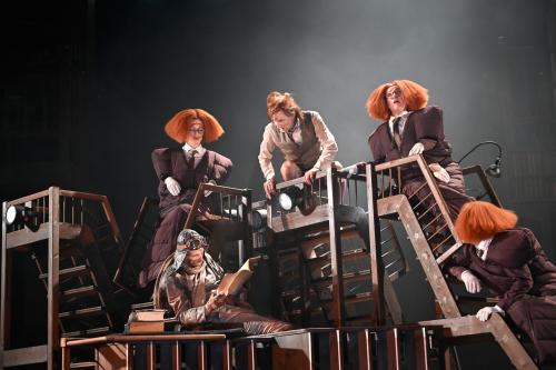 Auf einer Ansammlung von Treppen hockt Jan-Peter Kampwirth und blickt nach unten zu einer Person mit Pilotenmütze, die in einem Manuskript liest. Ebenfalls auf den Treppen sitzen drei Personen mit aufgeplusterten orangenen Perücken. Sie blicken in alle Richtungen.