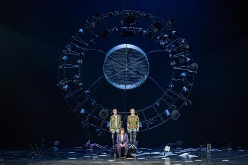 Eine senkrecht hängende große runde Stahlkonstruktion aus vielen unterschiedlich großen alten trichterförmigen Lautsprechern, Scheinwerfern und Ventilatoren. Davor stehen Kristof Van Boven und Nils Kahnwald auf einem Podest. Sie tragen schwarze Leggings mit weißen Mustern, kurze Jeanshosen darüber und dunkelgrüne Jacken mit Symbolen und Bildern, wie einem Spinnennetz, Sternen, Feuer, Herzen, Skeletten. Zu ihren Füßen sitzt Julia Wieninger in einem lila-grün karierten Anzug. Auf dem Boden verteilt liegen Pap