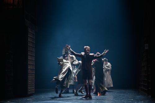 Jan-Peter Kampwirth steht mit ausgestreckten Armen und geöffnetem Mund im Vordergrund, hinter ihm sind mehrere Personen in Bewegung, die Trenchcoats tragen. Einer Frau fallen gerade Bücher aus der Hand.