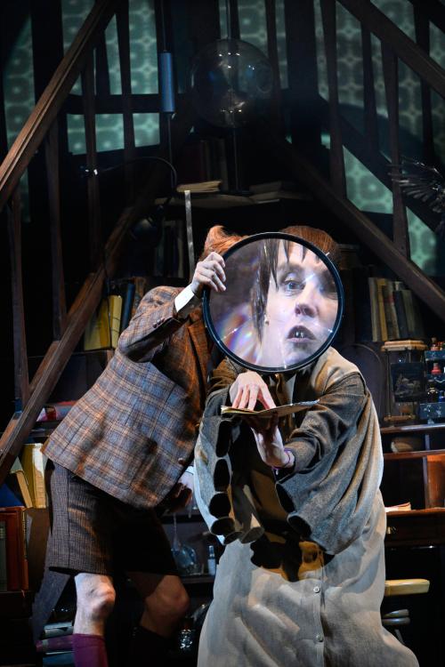 Jan-Peter Kampwirth und Ute Hannig stehen dicht nebeneinander und blicken durch ein großes Vergrößerungsglas, sodass vor allem Ute Hannigs Gesicht stark vergrößert ist. In den Händen hält sie ein Manuskript.