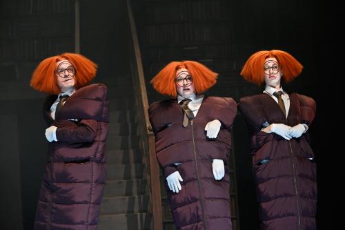 Drei Personen in Daunenjacken-Kleidern und mit orangen aufgeplusterten Perücken stehen nebeneinander vor einem großen Bücherregal. Die mittlere Person hat drei Hände. Sie blicken mit verschiedenen Gesichtsausdrücken in die Ferne.