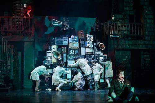 Aus mehreren Stereo- und Musikanlagen, Musikboxen und Grammophonen ist ein großer Gerätehaufen aufgebaut, vor dem fünf Personen stehen, die an den Anlagen rumdrehen. Jan-Peter Kampwirth sitzt im Schneidersitz mit geöffnetem Mund am Bühnenrand. Alberta von Poelnitz und Katja Danowski schauen von zwei Balkons im Hintergrund aus zu.