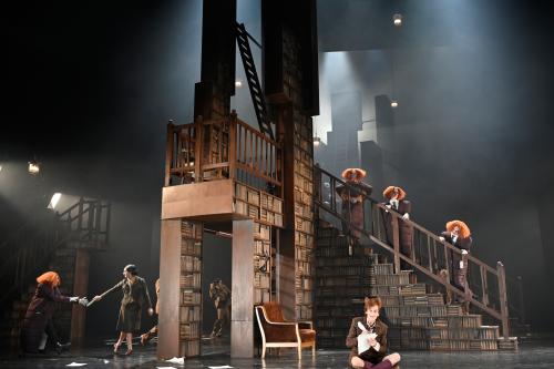 Drei Personen in Daunenjacken-Kleidern und mit orangen aufgeplusterten Perücken stehen nebeneinander auf einer Treppe eines großen Bücherregals. Im Hintergrund sind Tänzer*innen zu sehen. Jan-Peter Kampwirth sitzt im Schneidersitz mit geöffnetem Mund am Bühnenrand und schreibt etwas mit einer Feder auf ein Blatt Papier. 