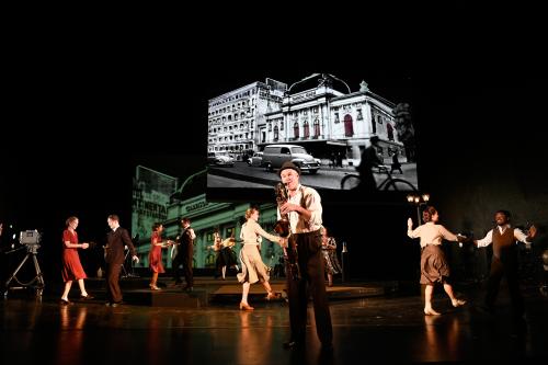 Bühnensituation: Yorck Dippe mit Hut, weißem Hemd und brauner Hose mit Hosenträgern und einem Saxofon in den Händen. Im Hintergrund verteilt sechs tanzende Paare, die sich an der Hand halten und je ein Bein nach vorn gestreckt. Über allem eine Leinwand mit einem schwarz-weißen Foto des SchauSpielHauses mit der Bezeichnung „Garrison Theatre“ mit alten Autos und Menschen auf Fahrrädern davor.