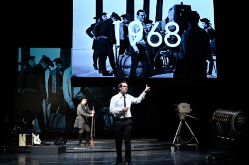 Bühnensituation: Daniel Hoevels mit Brille, weißem Hemd, Krawatte und Anzughose. Hinter ihm Matti Krause in Uniform und Dreispitz-Hut, der sich auf ein Gewehr stützt und zu Daniel Hoevels schaut. Dessen linke Hand weist auf die Leinwand über ihm, die ein schwarz-weißes Foto von mehreren Männern in Uniform und Dreispitz-Hut zeigt sowie einen Mann mit Brille, weißem Hemd, Krawatte und Anzughose, alle stehen vor einer alten Filmkamera. Über dem Bild liegt die Zahl 1968. Auch auf der Bühne ist eine alte Filmkam