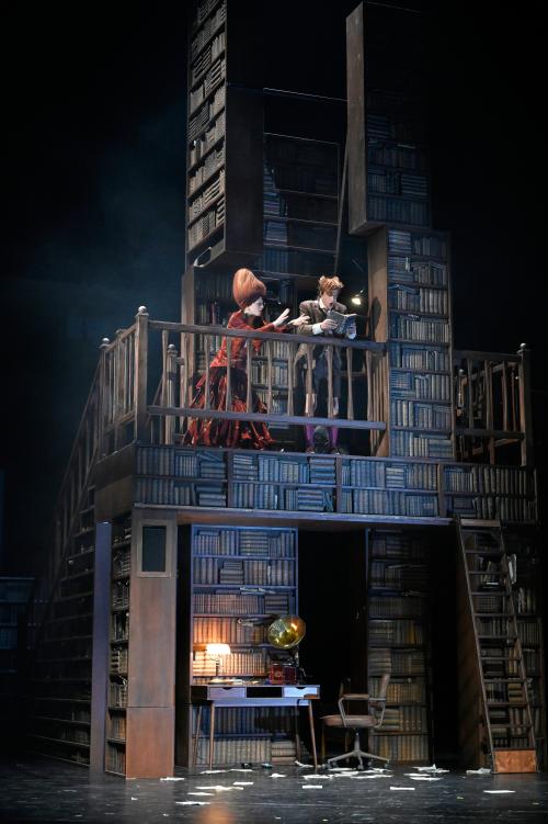 Auf einem Balkon inmitten vieler aufgetürmter Bücherregale stehen Alberta von Poelnitz und Jan-Peter Kampwirth. Er hat ein Buch aufgeschlagen und schaut mit geöffnetem Mund hinein, Alberta geht auf ihn zu und streckt die Arme nach dem Buch aus. 