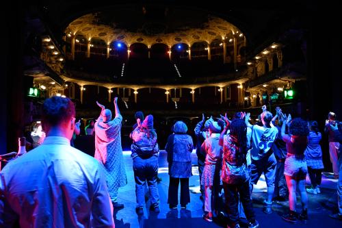 Bühnensituation, von der Bühne in den Saal fotografiert. Eine Gruppe von etwa zwanzig Menschen steht klatschend mit dem Rücken zur Kamera Richtung Saal. Sie werden von blauem und rotem Licht angestrahlt, im Hintergrund ist schwach beleuchtet der Zuschauerraum zu sehen.
