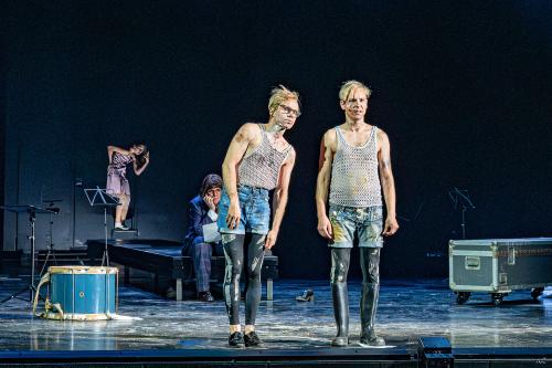 Kristof Van Boven und Nils Kahnwald stehen nebeneinander auf der Bühne. Kristofs Oberkörper neigt sich etwas in Richtung von Nils, die beiden blicken in dieselbe Richtung. Im Hintergrund sitzt Julia Wieninger auf einem Podest und beobachtet die beiden. Auf der Bühne verstreut stehen Notenständer, eine Kiste und eine Trommel. Weiter im Hintergrund tanzt eine Tänzerin.