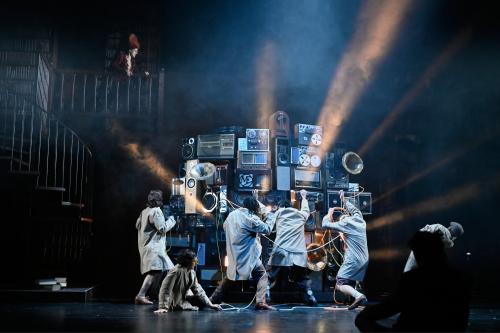 Aus mehreren Stereo- und Musikanlagen, Musikboxen und Grammophonen ist ein großer Gerätehaufen aufgebaut, vor dem fünf Personen stehen. Manche strecken die Arme nach dem Komplex aus, andere kämpfen mit Kabeln, die daraus kommen. Alberta von Poelnitz schaut von einem Balkon aus zu.