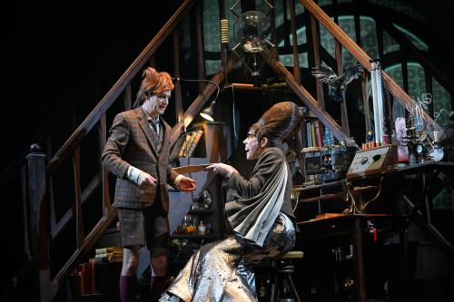 Jan-Peter Kampwirth steht vor zwei gegenüber zulaufenden Treppen in einem eingerichteten Büro, mit vielen wissenschaftlichen Instrumenten. Neben ihm sitzt Ute Hannig auf einem Stuhl und hält ihm eine Mappe hin. 