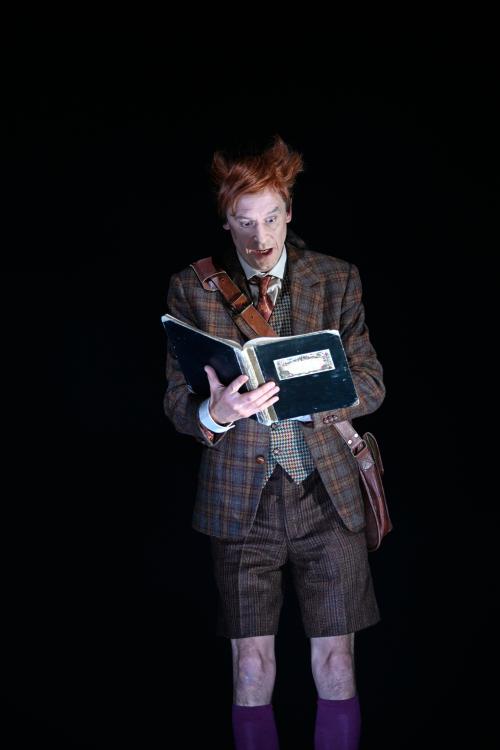 Jan-Peter Kampwirth steht alleine auf der dunklen Bühne. In den Händen hält er ein Buch, in das er mit geöffnetem Mund und weit aufgerissenen Augen schaut. Er trägt einen braun karierten Anzug mit kurzer Hose.