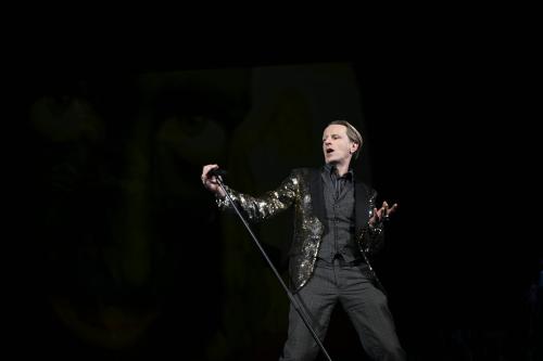 Bühnensituation: Alexander Scheer mit einem grau-karierten Anzug sowie einem gold-funkelnden Pailletten-Jackett. Er hält ein Mikrofon auf einem Ständer in seiner rechten Hand und kippt den Ständer zur Seite. Seine rechte Hand weist nach vorn, die Finger sind gespreizt. Er schaut zur Seite, sein Mund ist geöffnet.