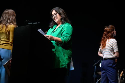 Bühnensituation: Eva Mattes in einer grünen Bluse an einem schwarzen Stehpult. Sie hält einige Blätter Papier in der Hand und schaut auf sie herab. Im Hintergrund sind zwei Personen von hinten zu sehen, die gerade stehen.